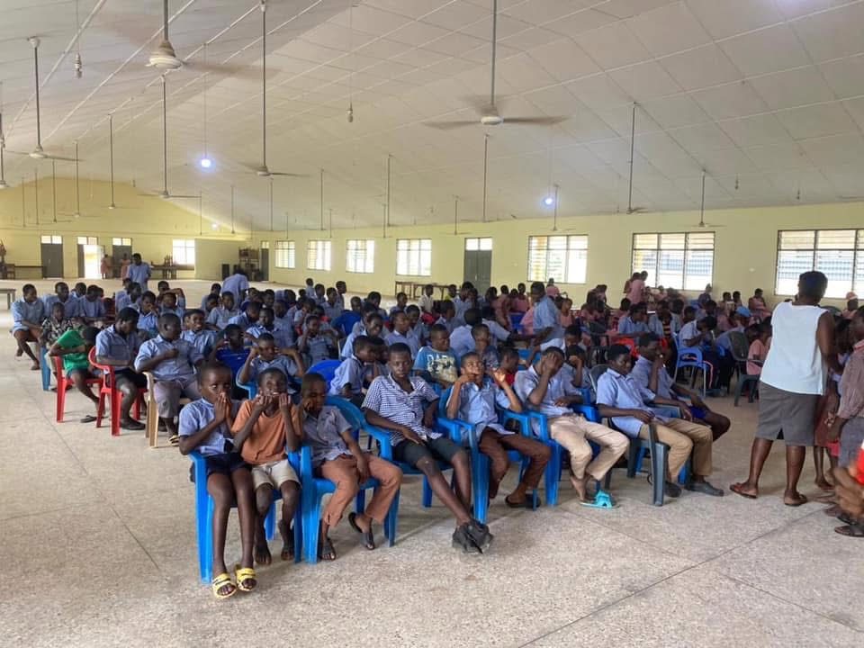 Rows of students in school uniform seated on plastic chairs in a large hall.